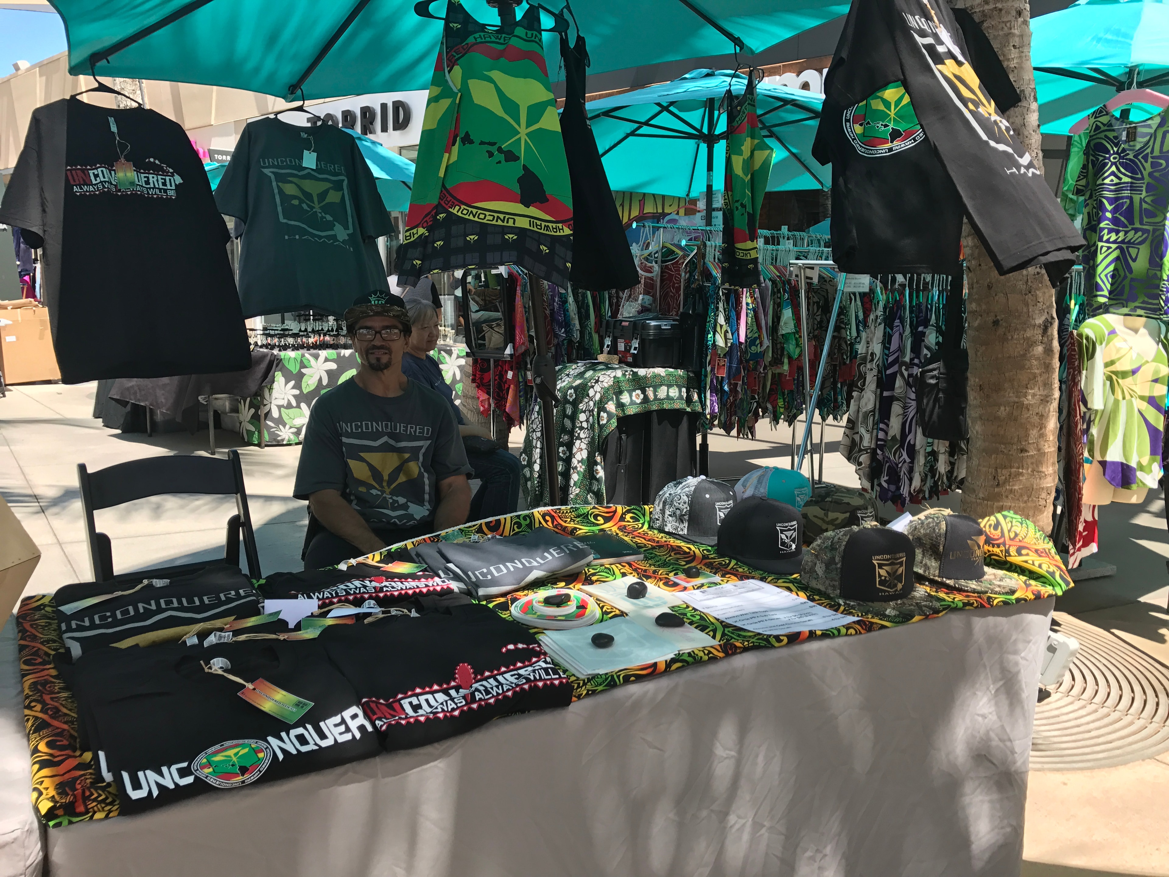 Unconquered Hawaii vendor booth with branded t-shirts, hats, and accessories at outdoor market
