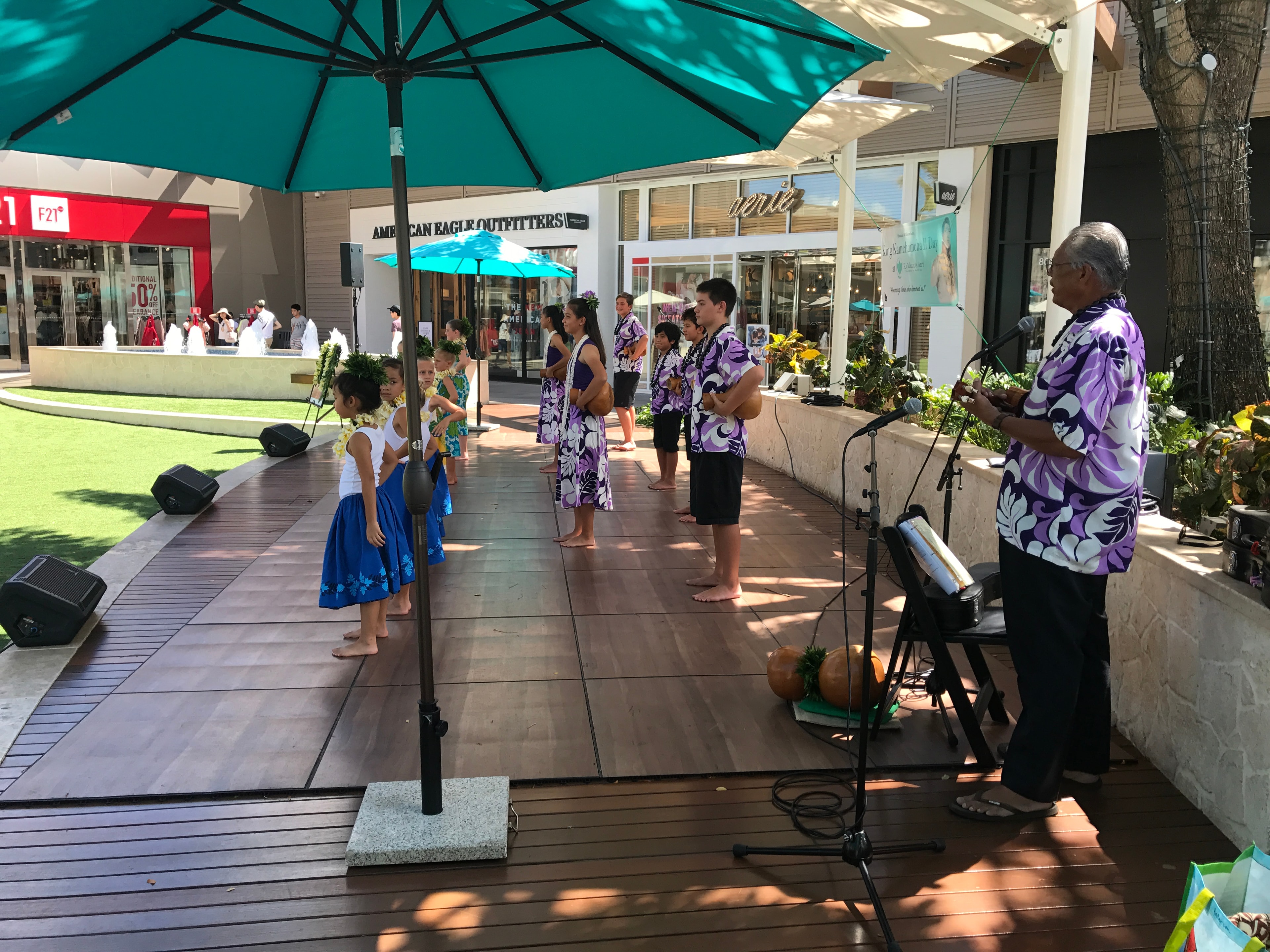 Hula halau lined up on outdoor stage with musician at King Kamehameha II Day at Ka Makana Alii