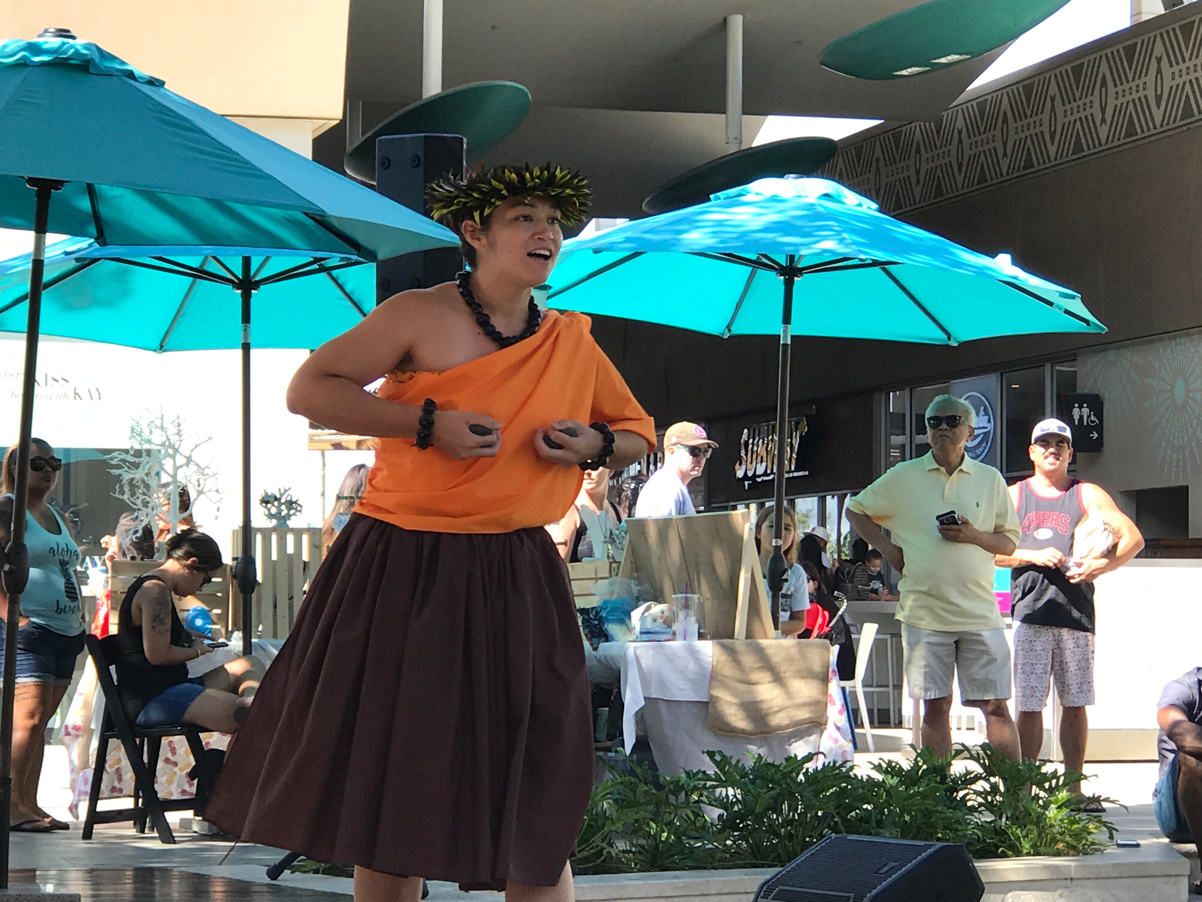 Hula dancer in orange kapa performing with crowd watching at Ka Makana Alii outdoor stage