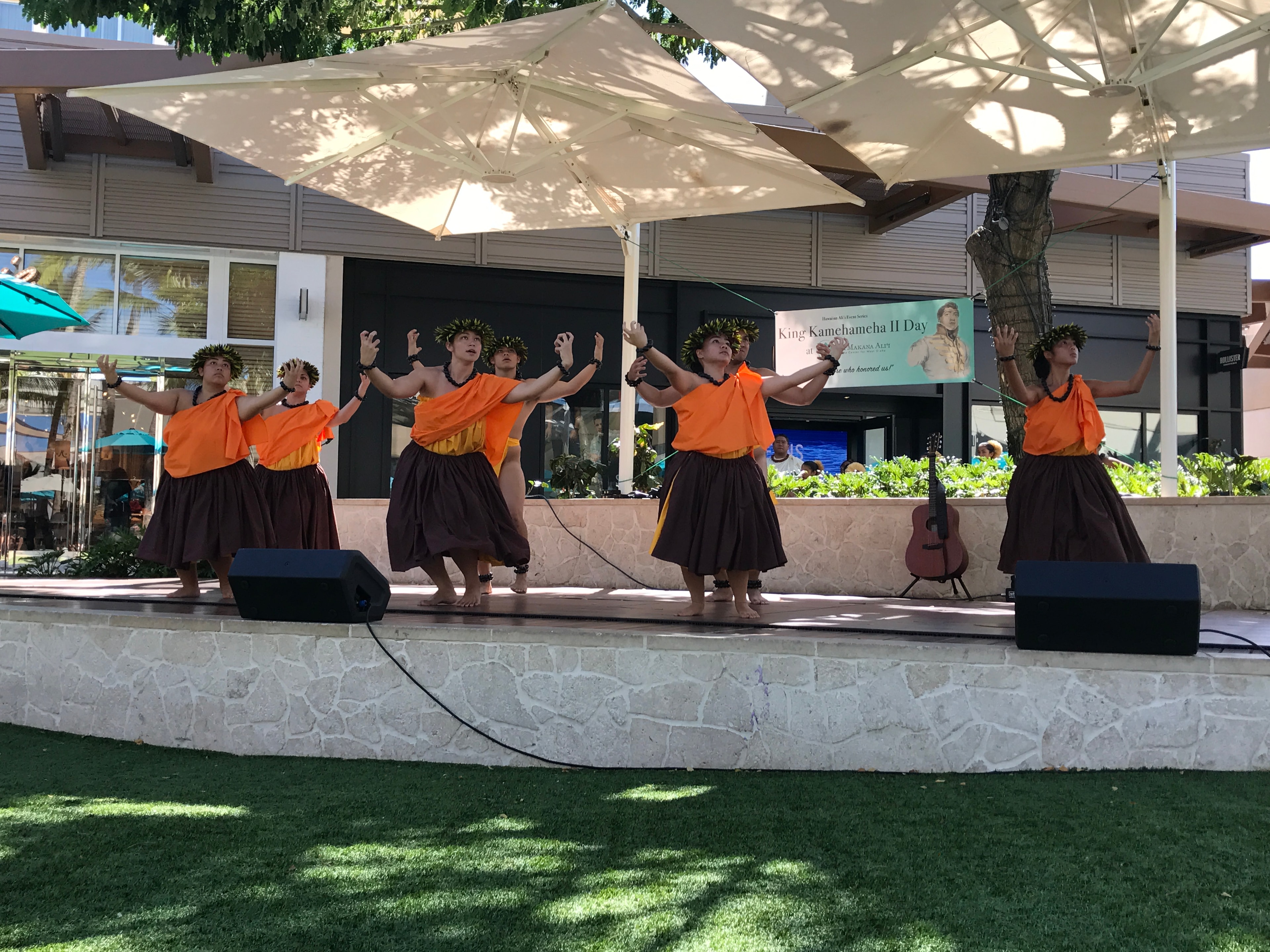 Hula halau in orange and brown performing with arms raised at King Kamehameha II Day stage