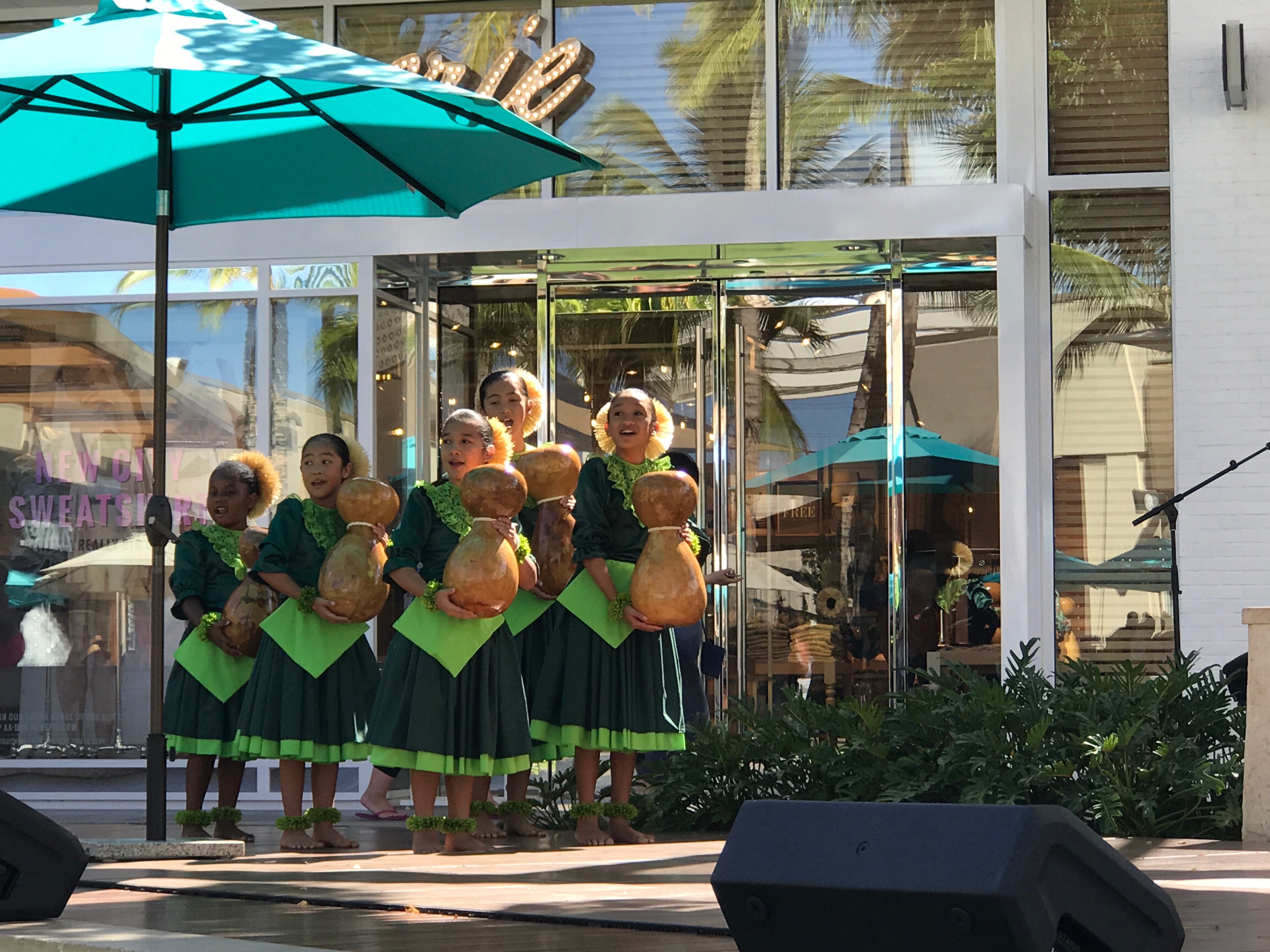 Keiki hula dancers in green dresses holding ipu gourds singing at outdoor mall stage