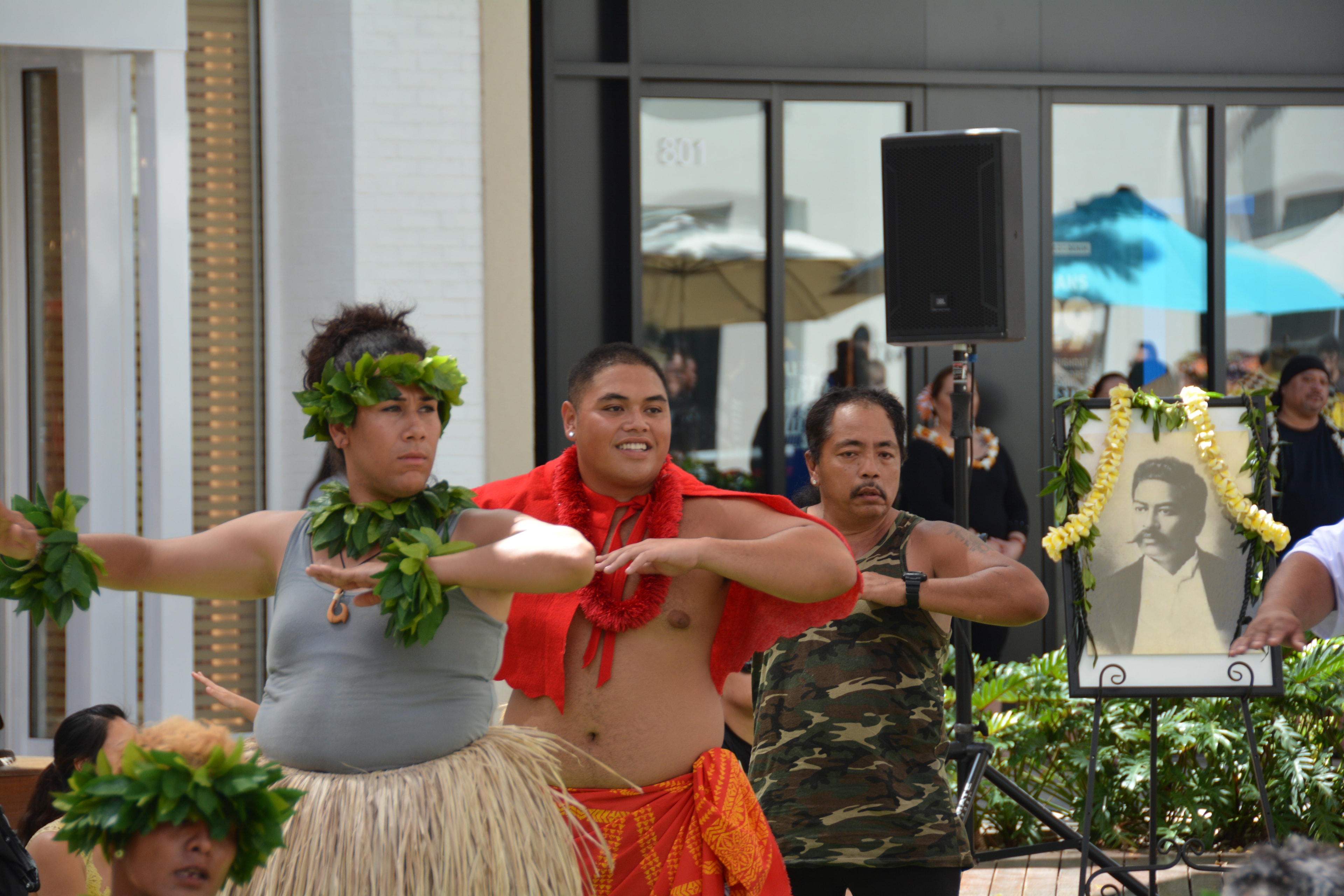 Hawaiian performers dancing with portrait of Prince Kūhiō in background