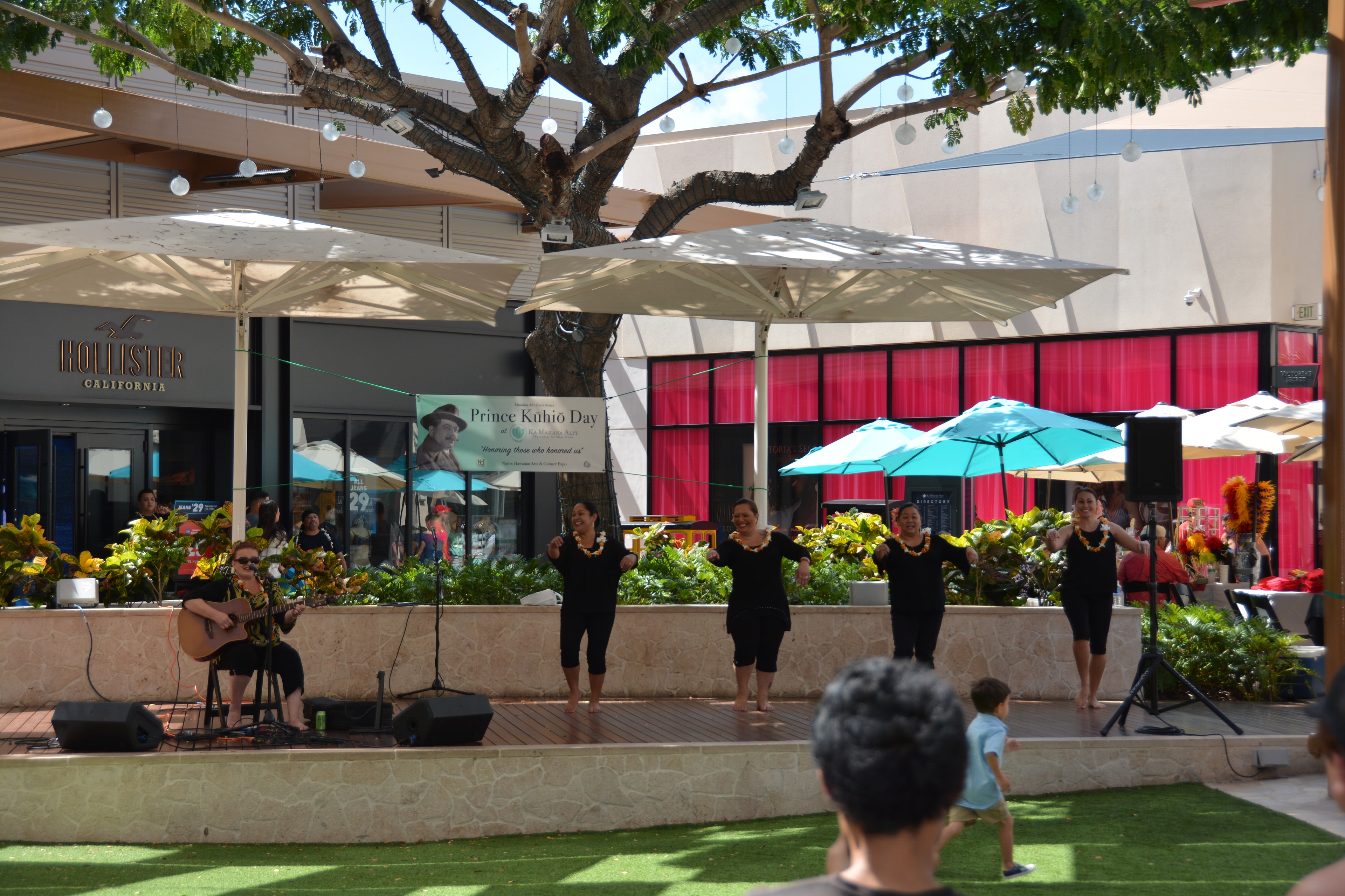 Hawaiian women dancers with orange and white lei performing at Prince Kūhiō Day stage