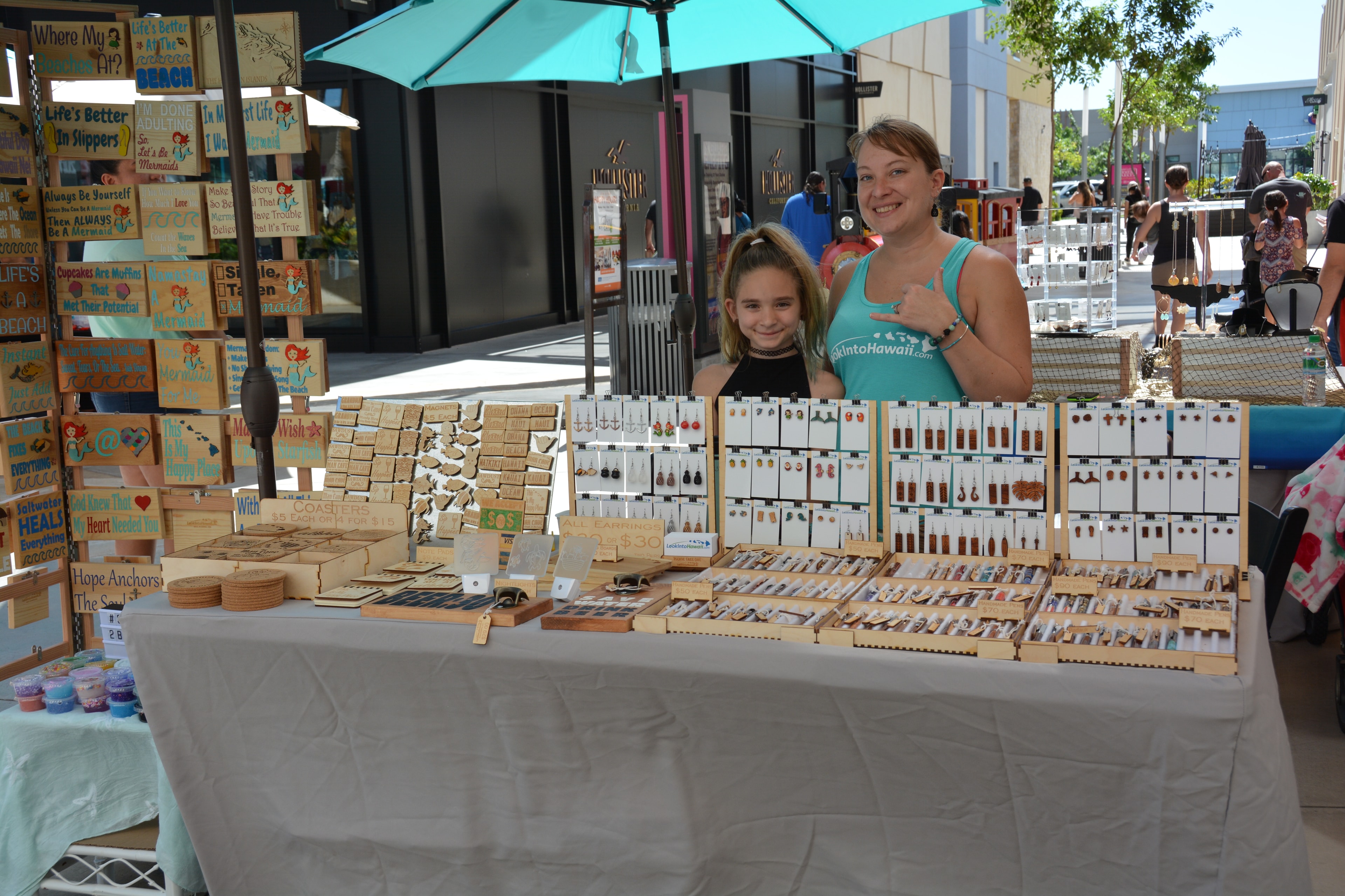Woman and girl at LookIntoHawaii vendor booth with wooden earrings and crafts at Prince Kūhiō Day market