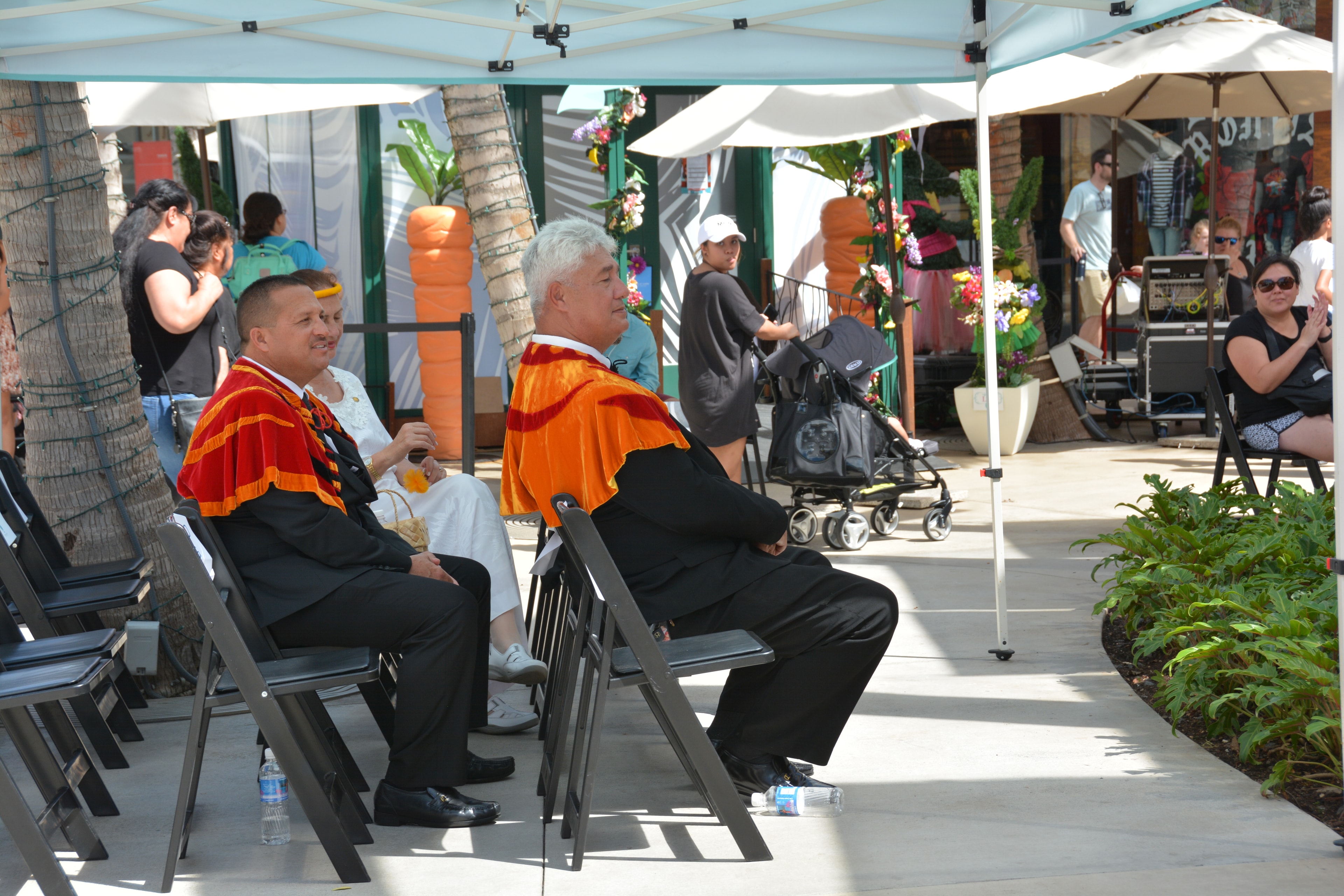 Two men in orange ceremonial robes seated at outdoor Prince Kūhiō Day celebration