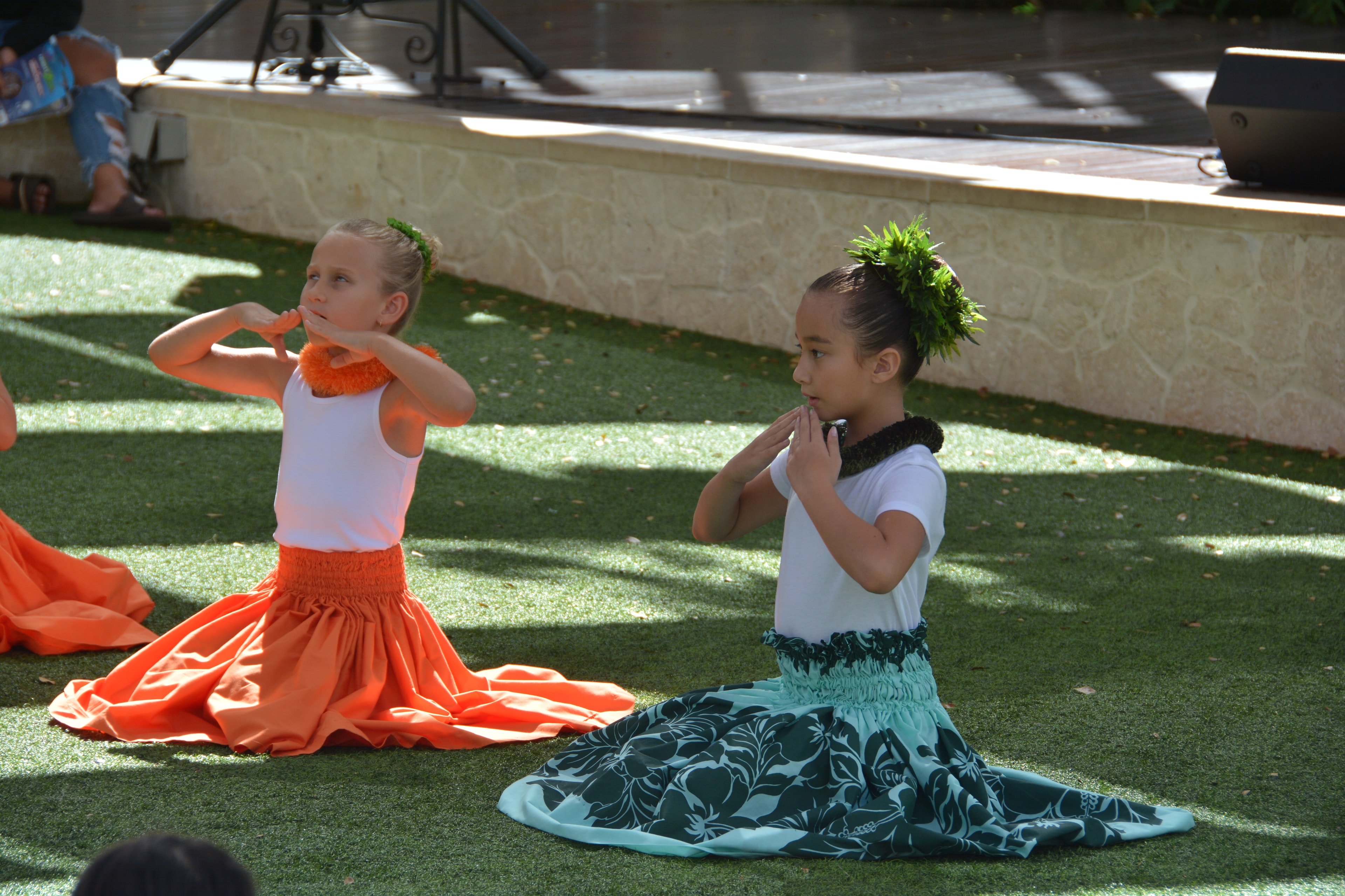 Two young girls in orange and teal hula skirts performing seated hula on artificial turf