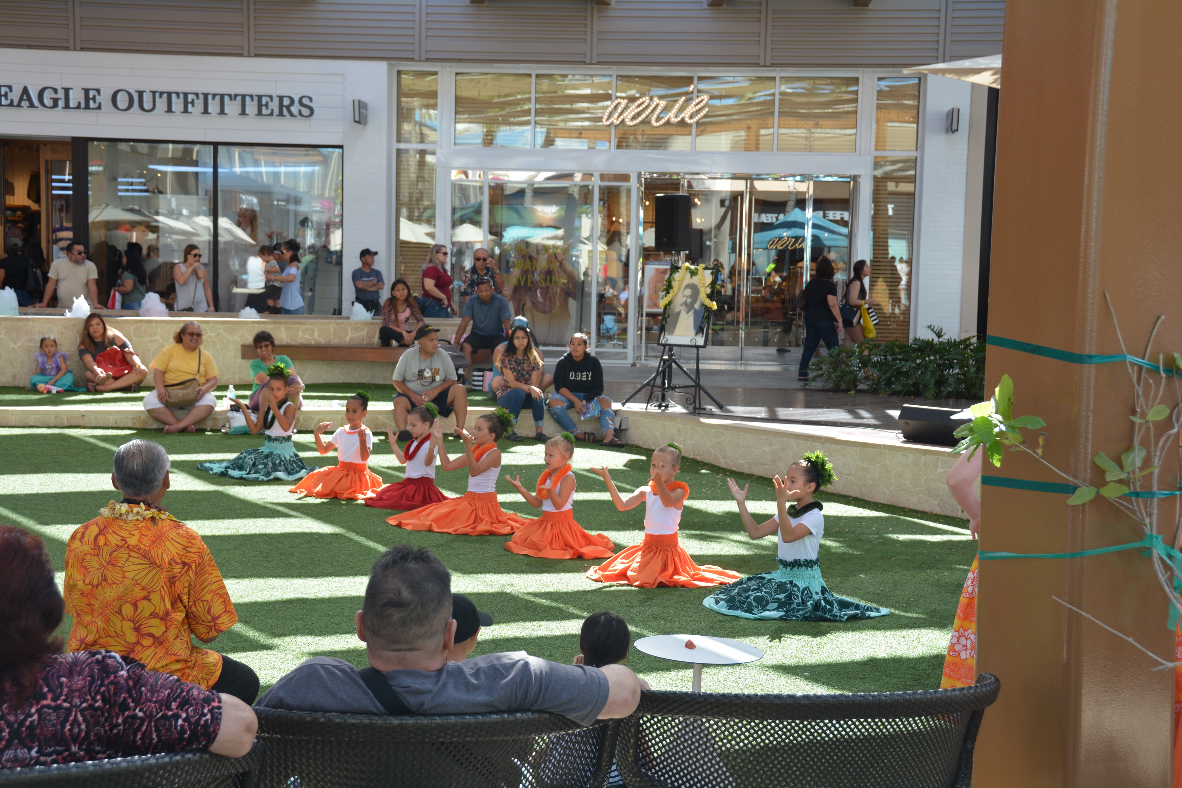 Children in orange hula skirts performing seated hula with audience watching at Ka Makana Aliʻi