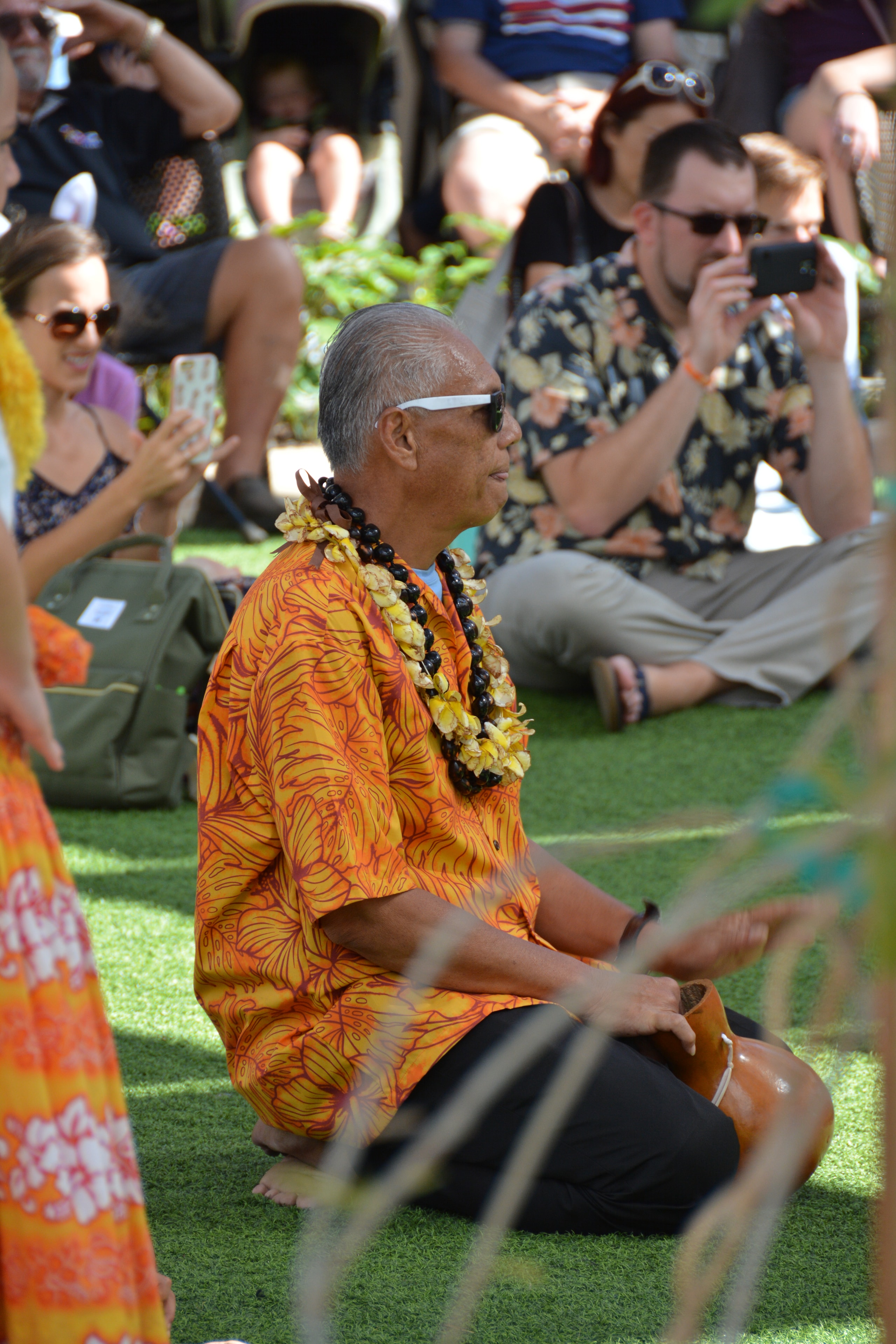 Elder man in orange floral Hawaiian shirt with lei watching hula performance at Prince Kūhiō Day