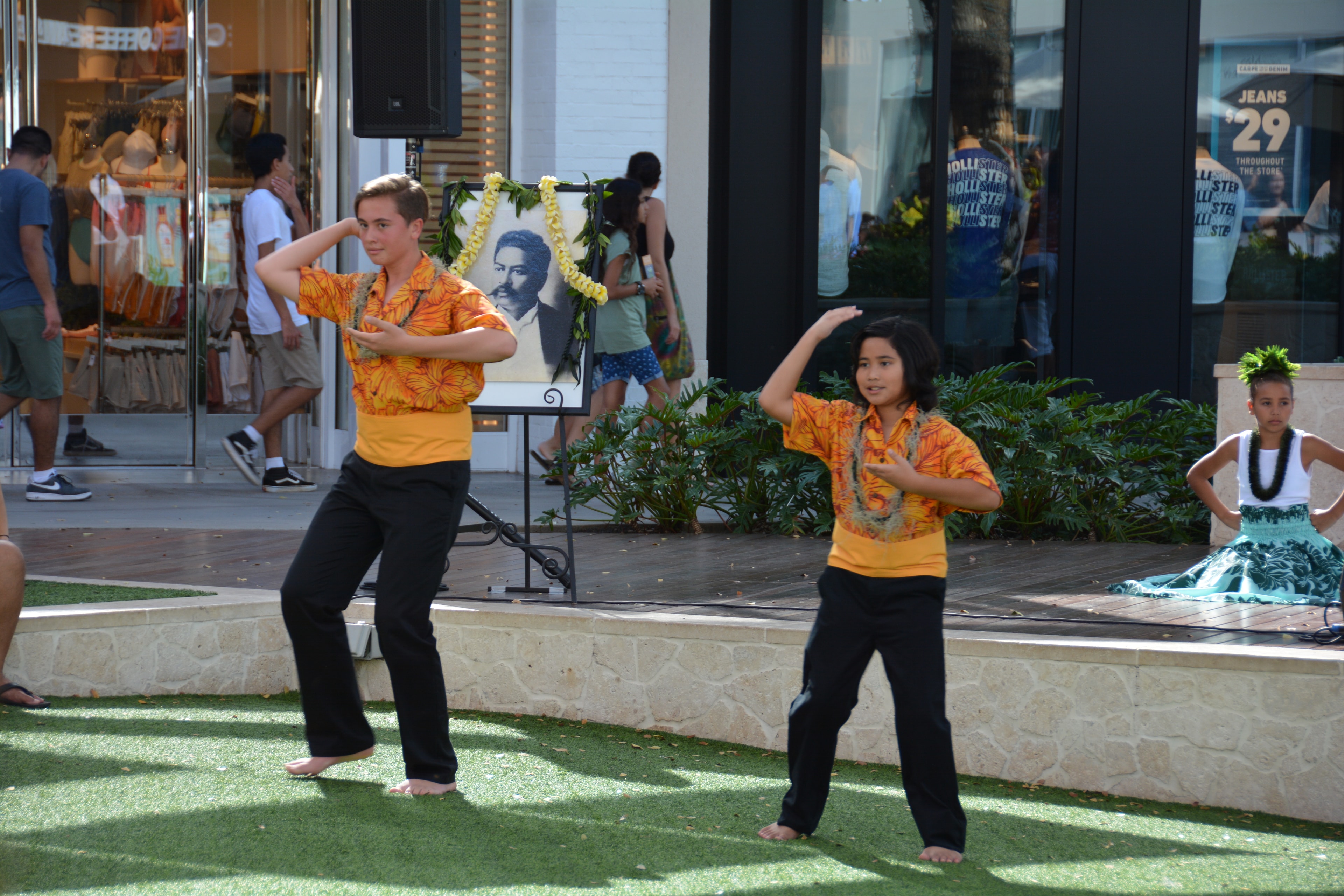 Two young male hula dancers in orange shirts performing with portrait of Prince Kūhiō in background