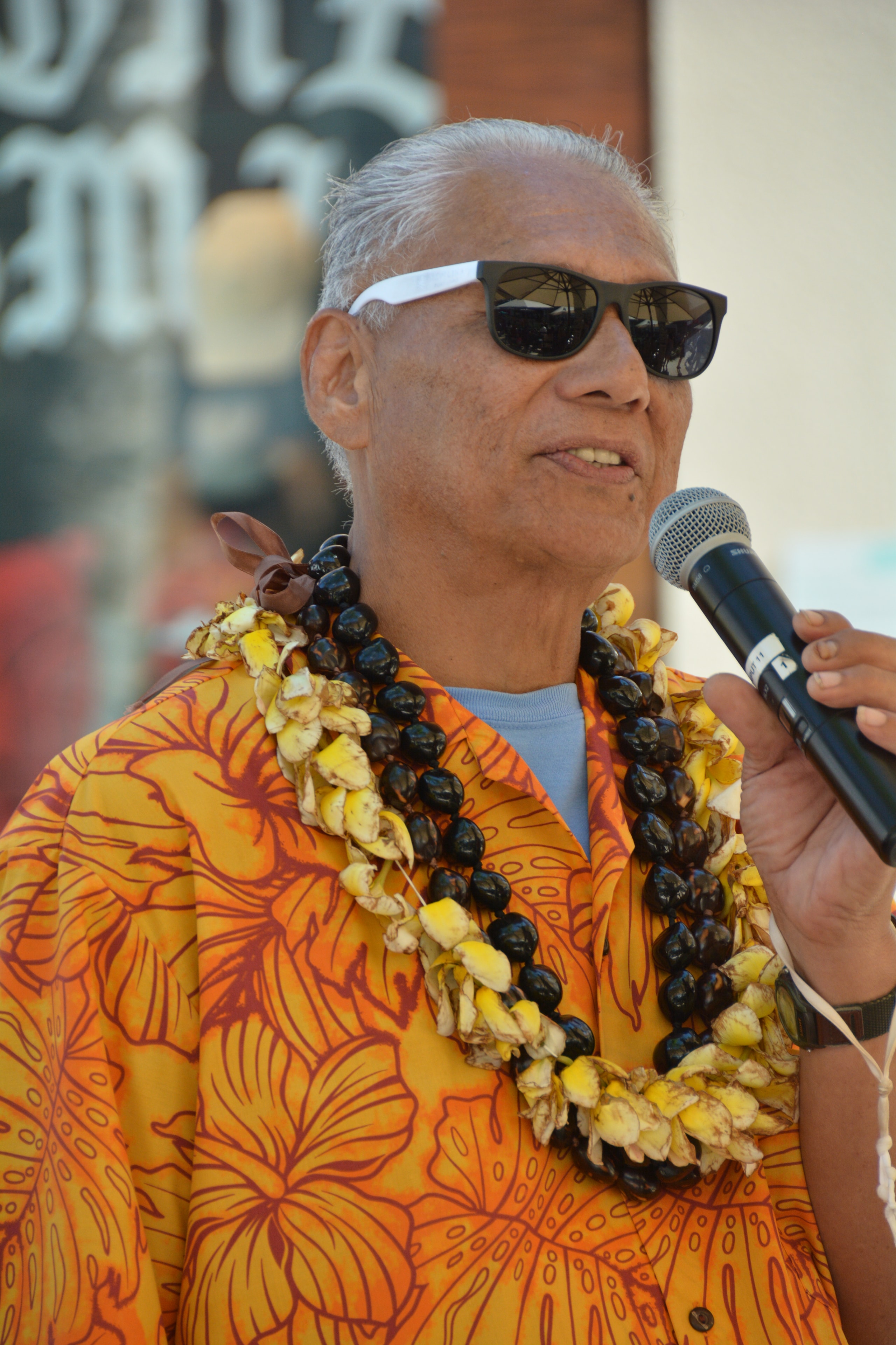 Elder man in orange floral shirt and black kukui nut lei speaking into microphone at Prince Kūhiō Day