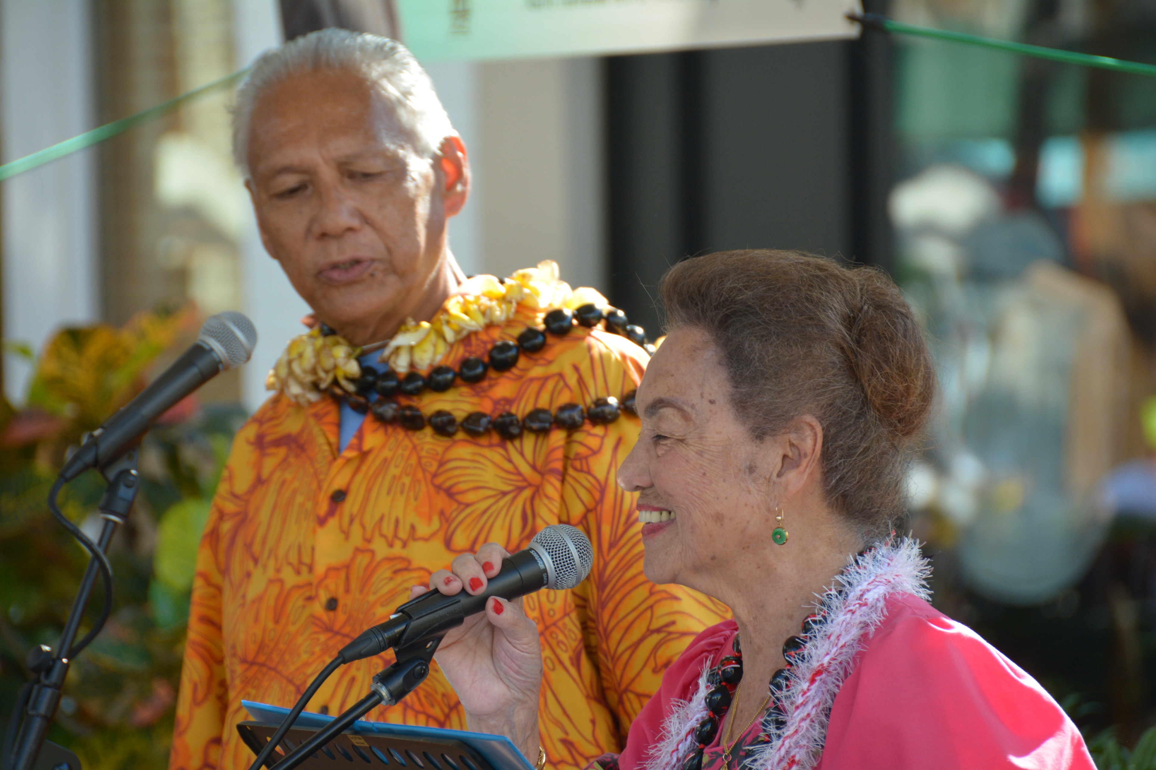 Man in orange floral shirt and woman in pink muʻumuʻu at microphone stand at Prince Kūhiō Day ceremony