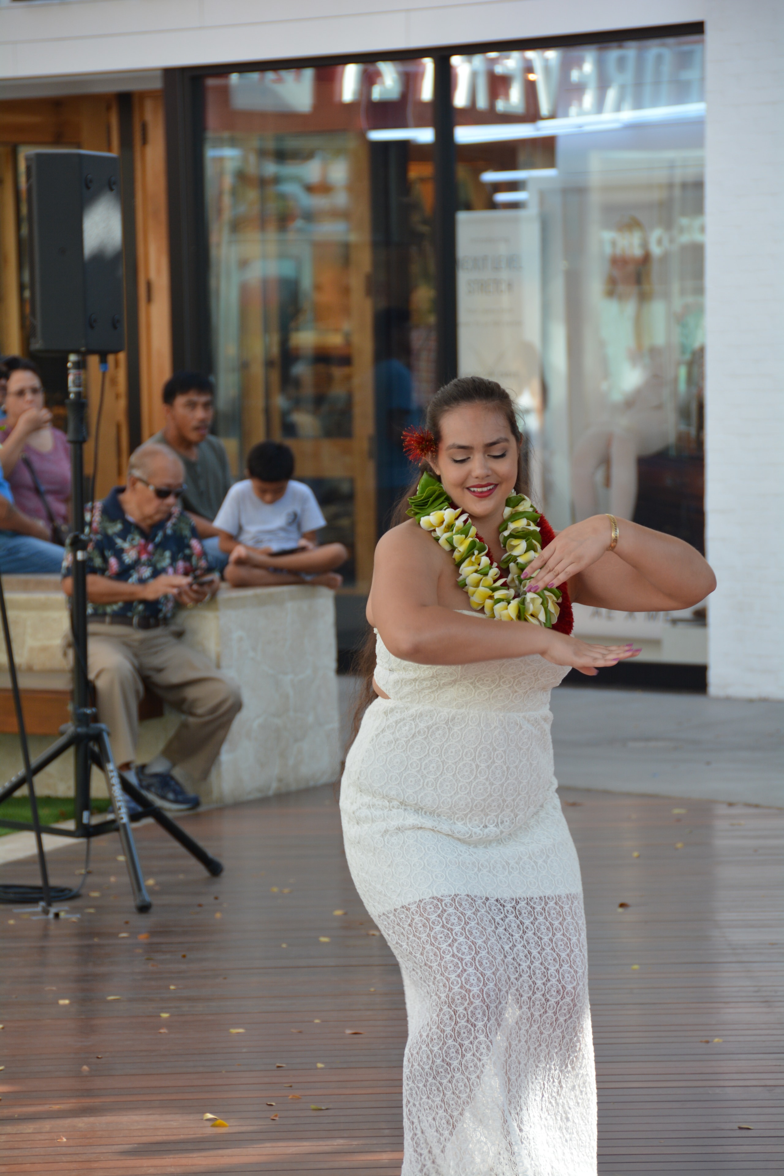 Woman in white lace dress with plumeria and red lei performing hula at outdoor Ka Makana Aliʻi stage
