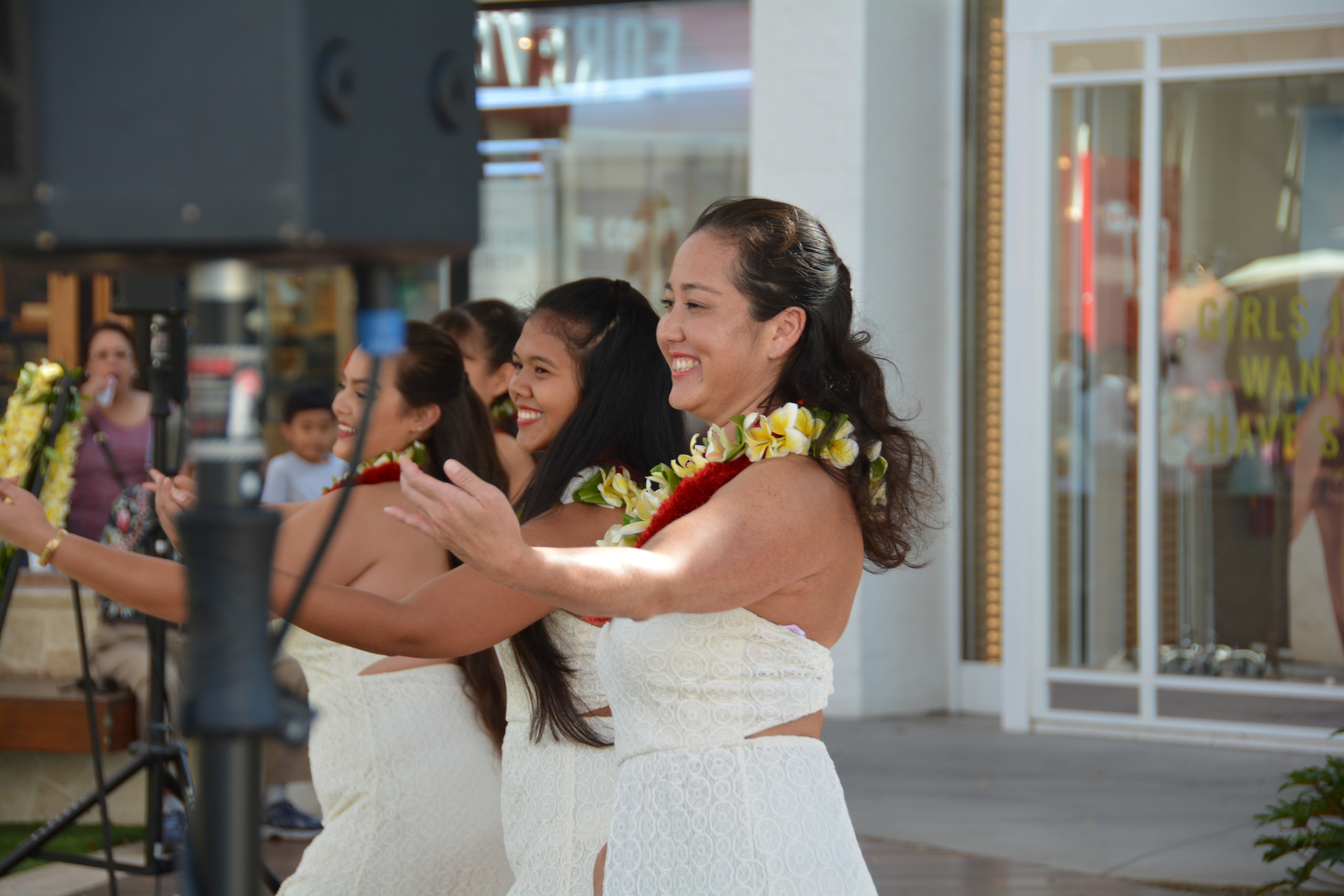 Group of Hawaiian women in white lace dresses with red and yellow lei smiling while performing hula