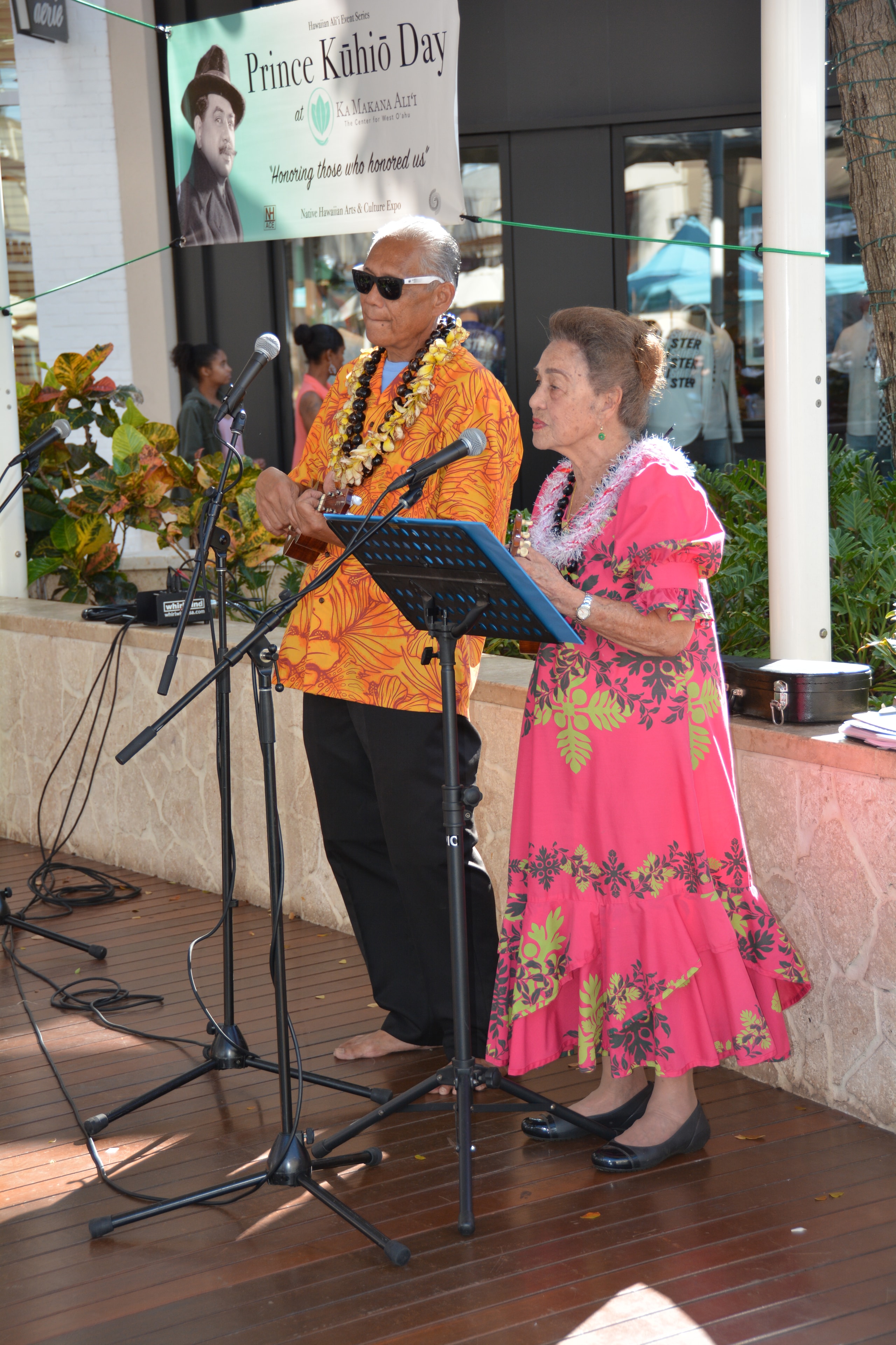 Elder man in orange floral shirt playing ukulele with woman in pink muʻumuʻu at Prince Kūhiō Day stage under banner