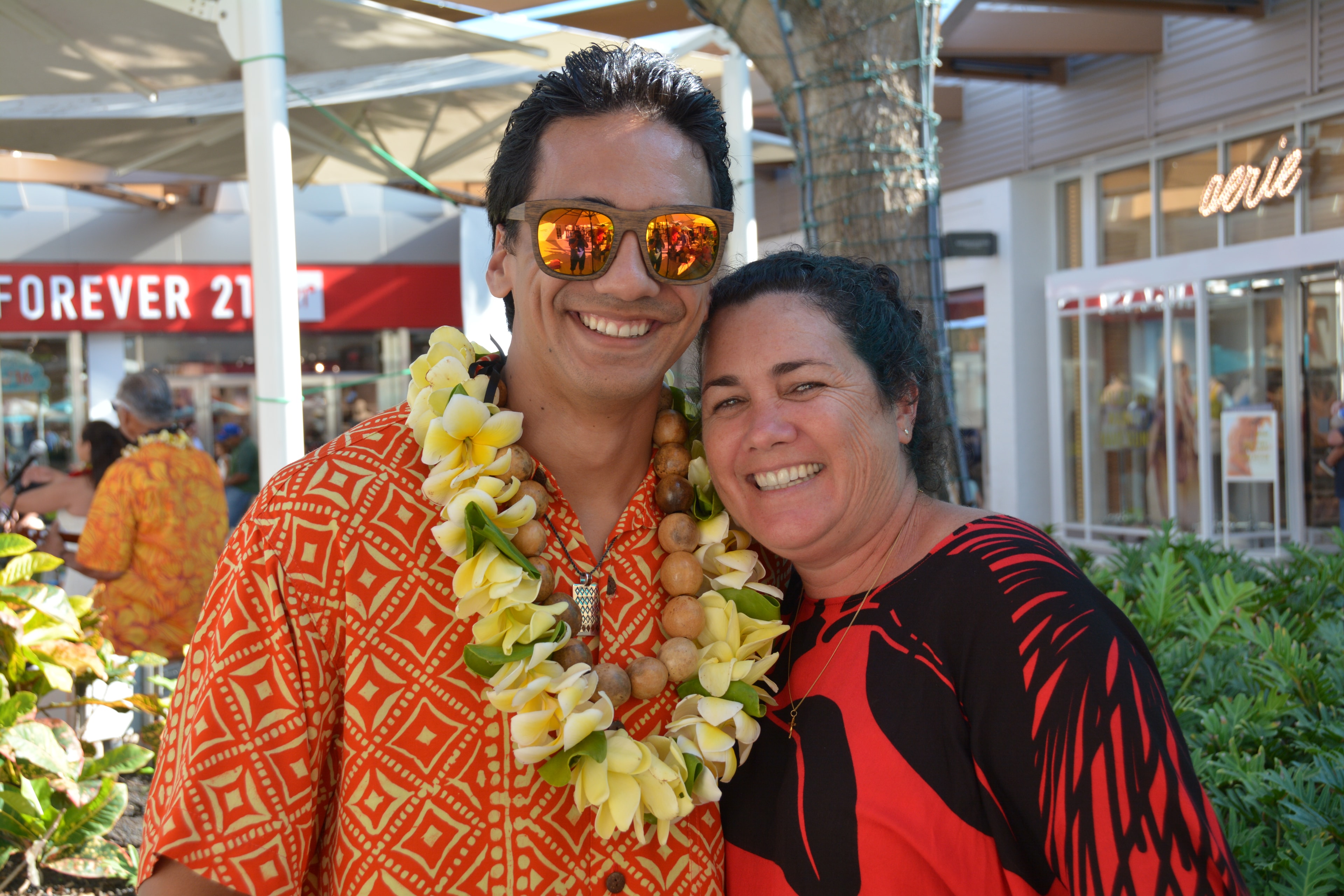 Young man in orange patterned Hawaiian shirt with plumeria and kukui nut lei smiling with woman at Ka Makana Aliʻi