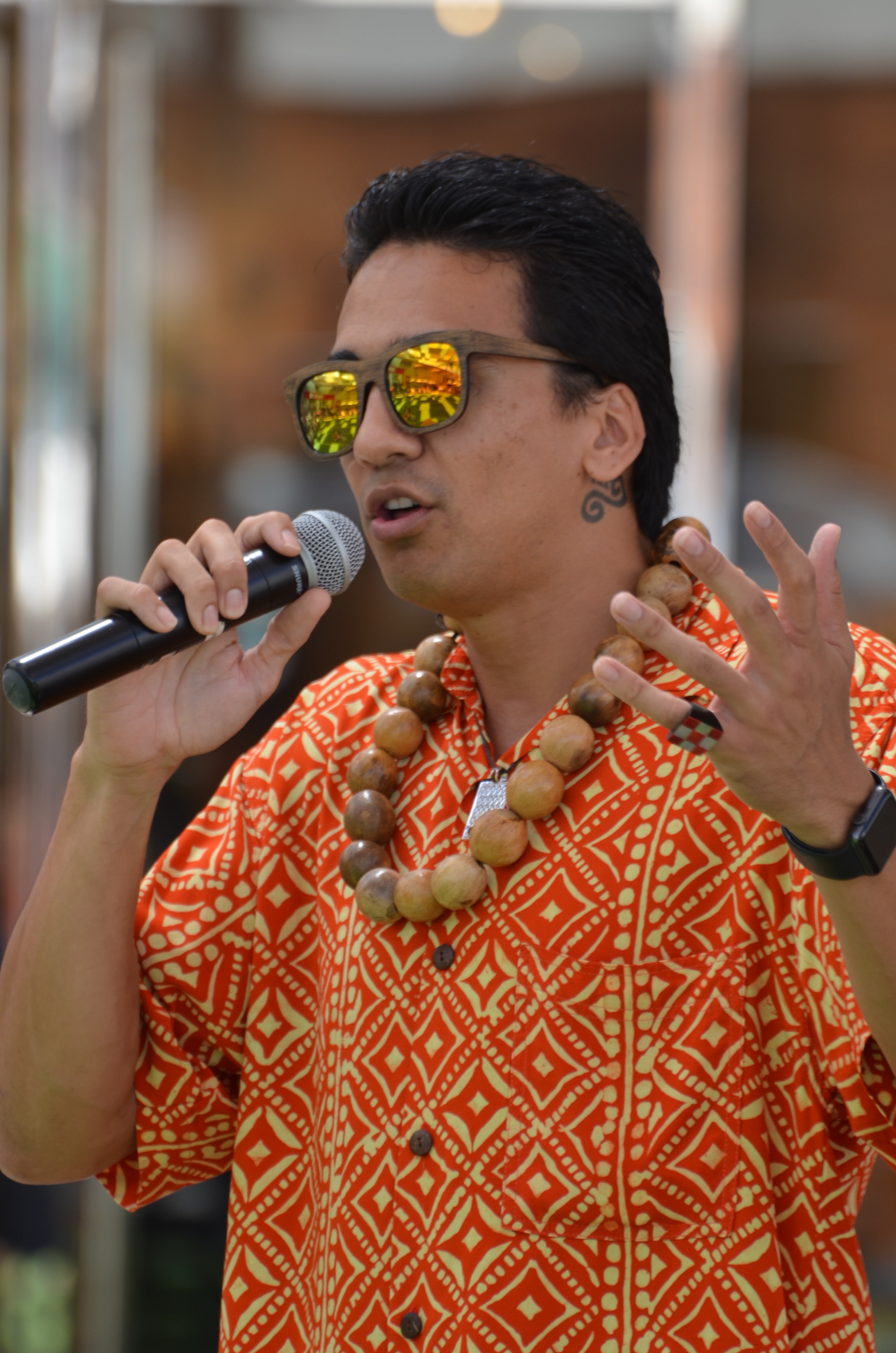 Man in orange patterned Hawaiian shirt with kukui nut lei speaking into microphone at Prince Kūhiō Day