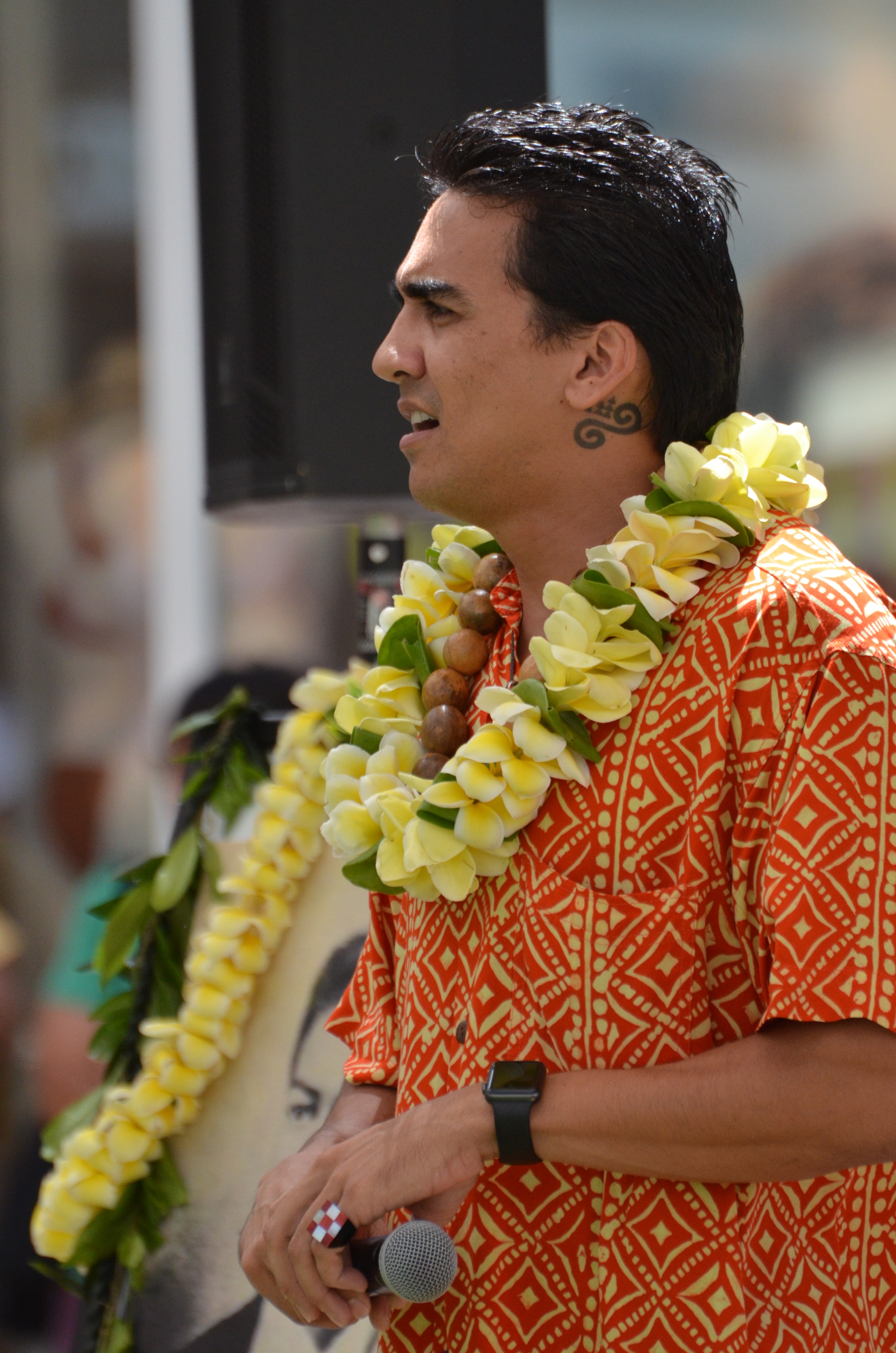 Man in orange patterned shirt with plumeria lei in profile holding microphone at Prince Kūhiō Day ceremony