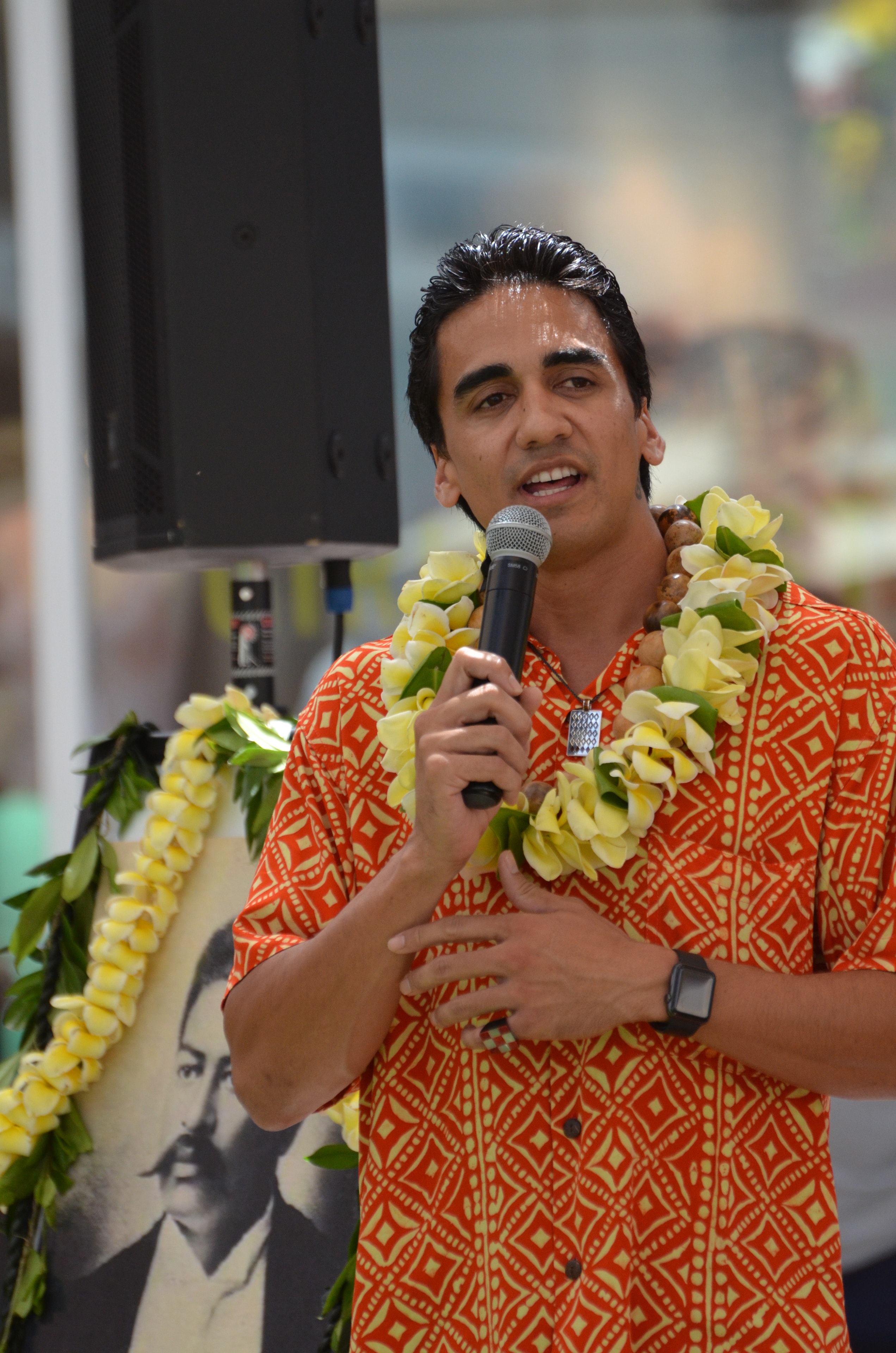 Man in orange patterned shirt with plumeria lei speaking into microphone at Prince Kūhiō Day with portrait of Prince Kūhiō behind him