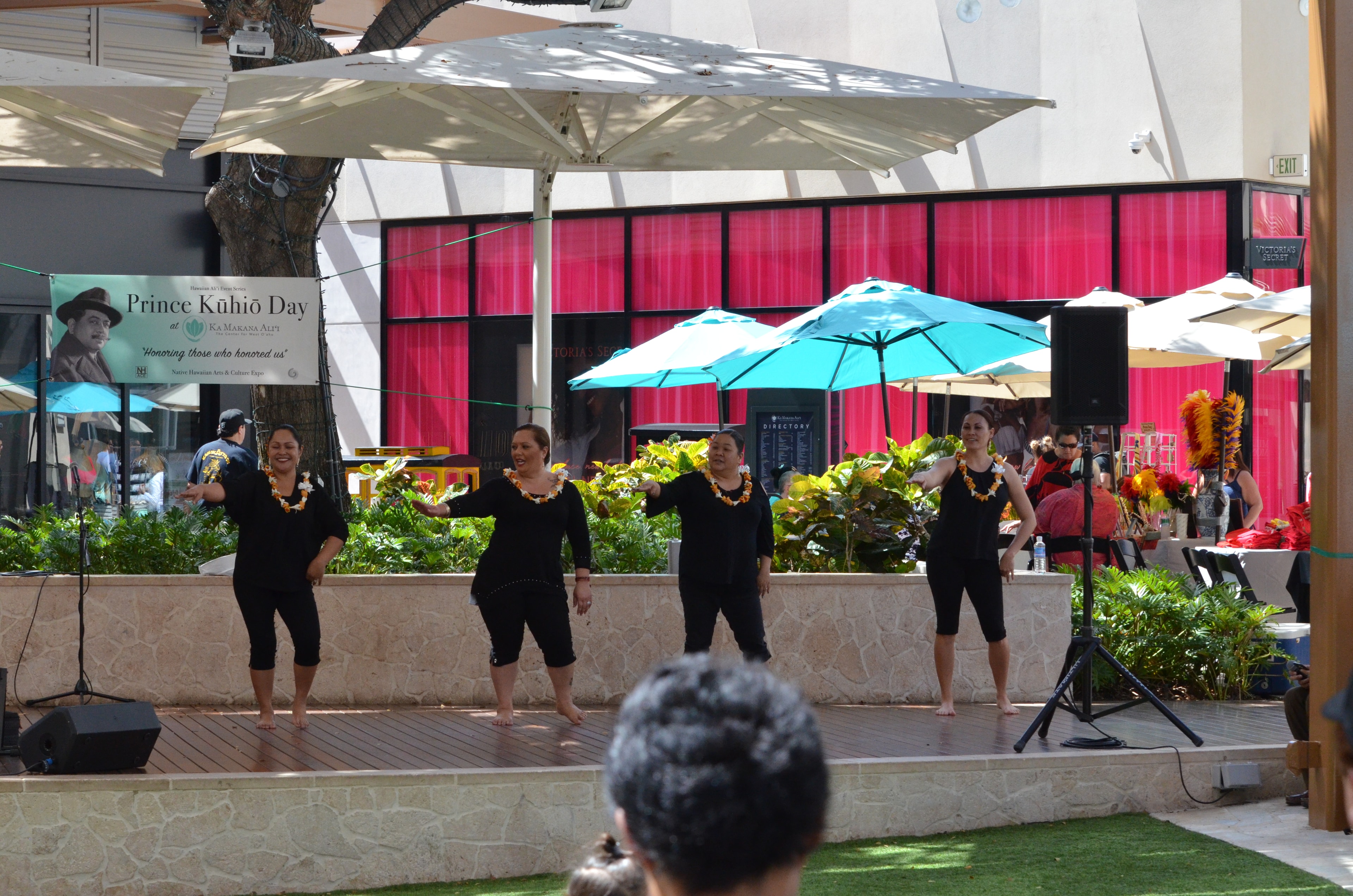 Four women in black with orange and white lei performing hula on stage at Prince Kūhiō Day at Ka Makana Aliʻi with Prince Kūhiō Day banner behind them