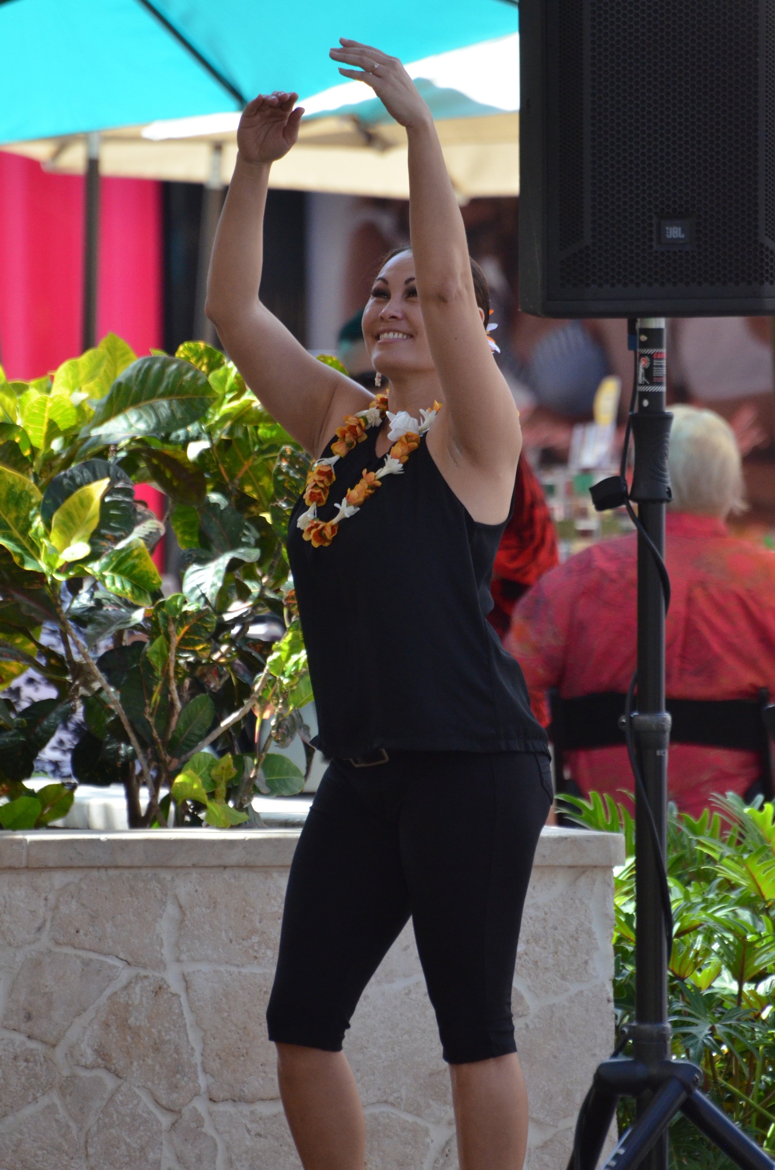 Woman in black with orange and white lei smiling with arms raised performing hula at Prince Kūhiō Day outdoor stage