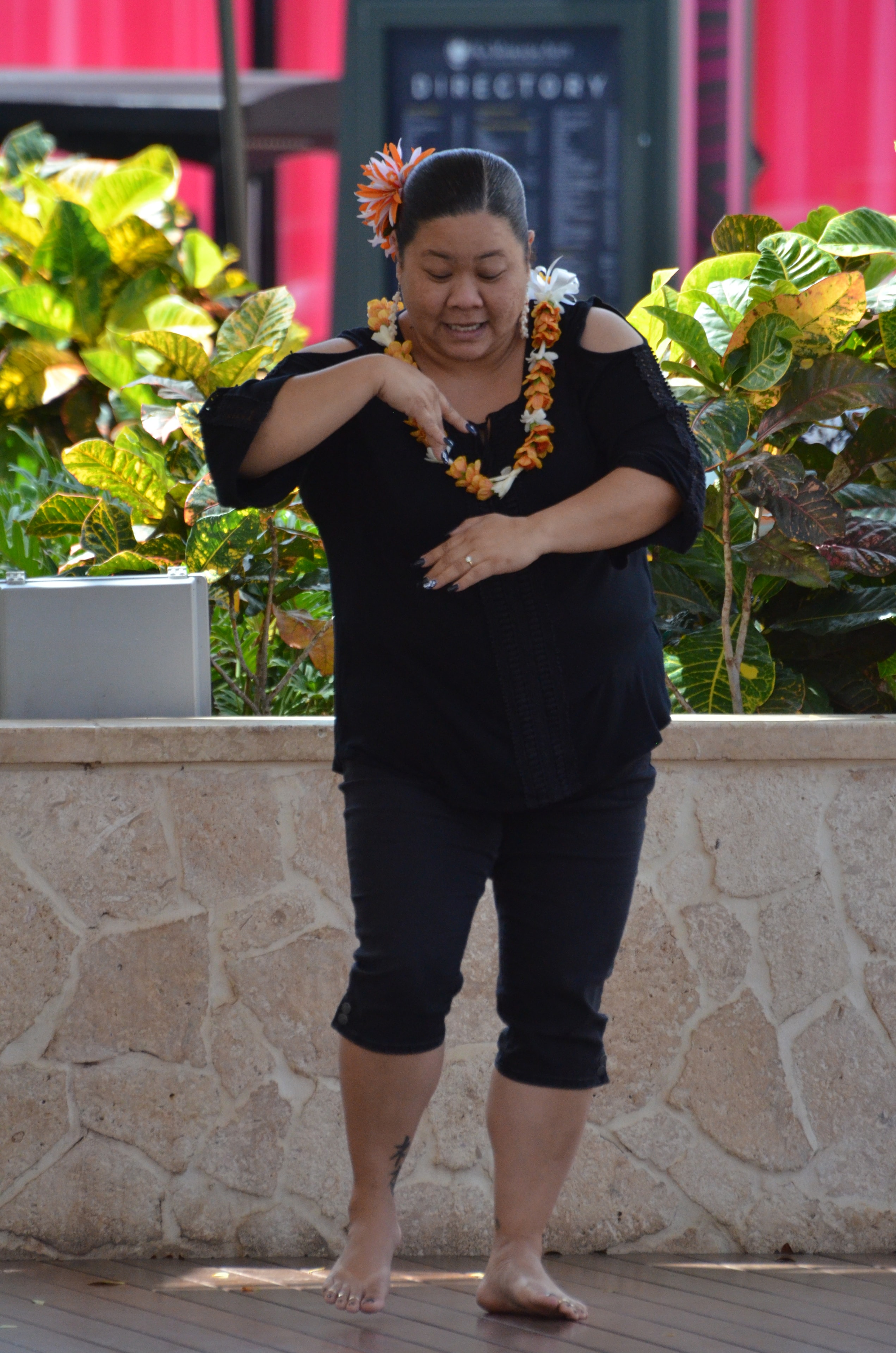 Woman in black with orange flower in hair and lei performing hula barefoot at Ka Makana Aliʻi stage