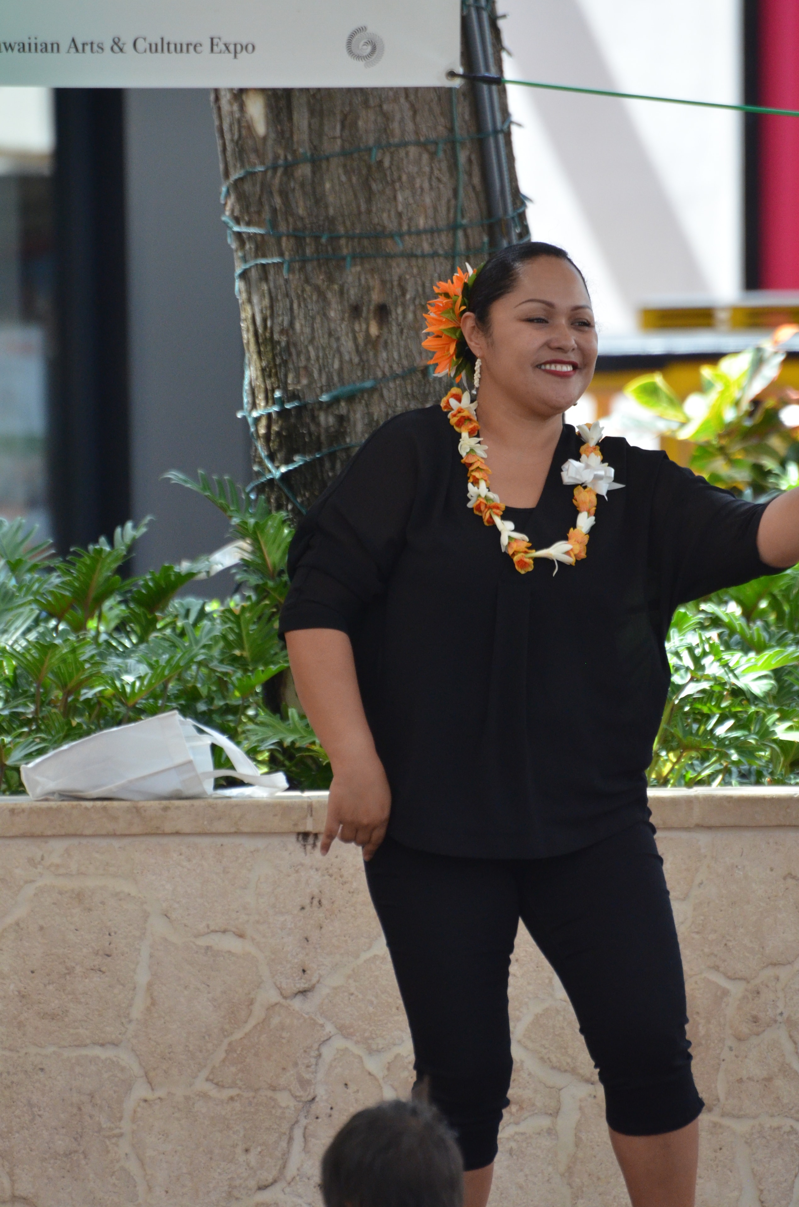Smiling woman in black with orange sunflower in hair and orange and white lei performing hula at Prince Kūhiō Day